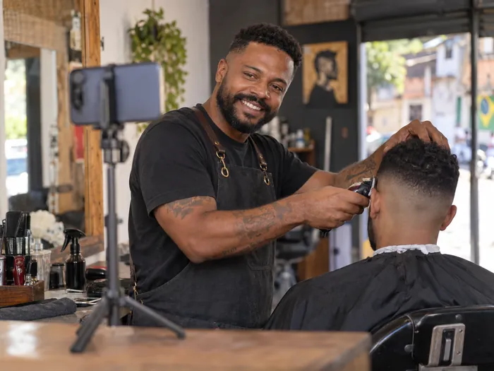 Barbeiro gravando com o celular num tripé enquanto corta o cabelo de um cliente